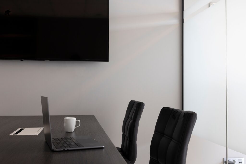 A sleek, modern office setup with a laptop, cup, and TV in an empty conference room.